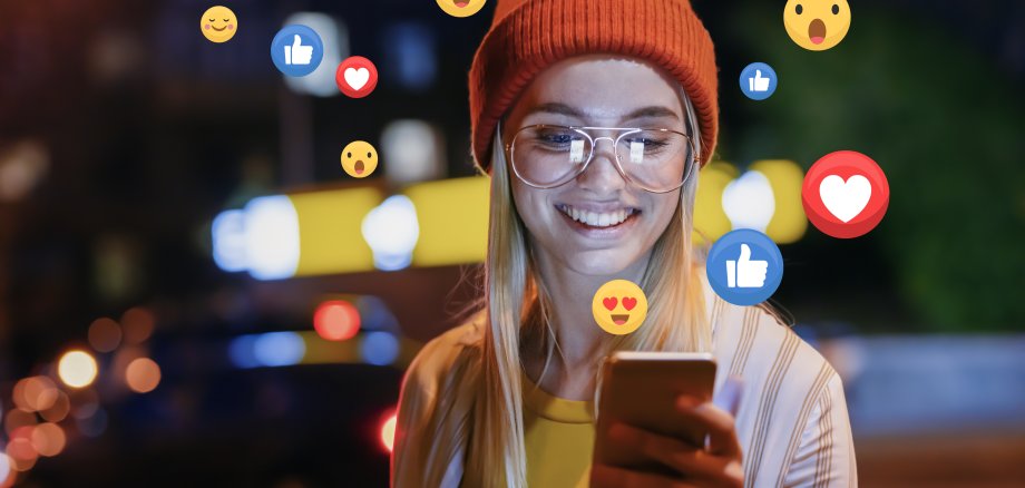 Junge Frau hält ein Smartphone in der Hand. Aus dem Smartphone strömen Emojis und Emoticons.