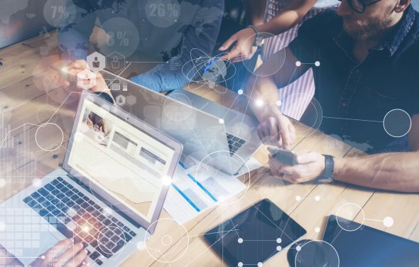 Vier Personen arbeiten zusammen. Hierbei rufen zwei Informationen über das Smartphone ab, zwei Personen arbeiten am Laptop. Zudem liegen noch zwei Tablets bereit. Im Hintergrund sieht man digitale Grafiken und Symbole.