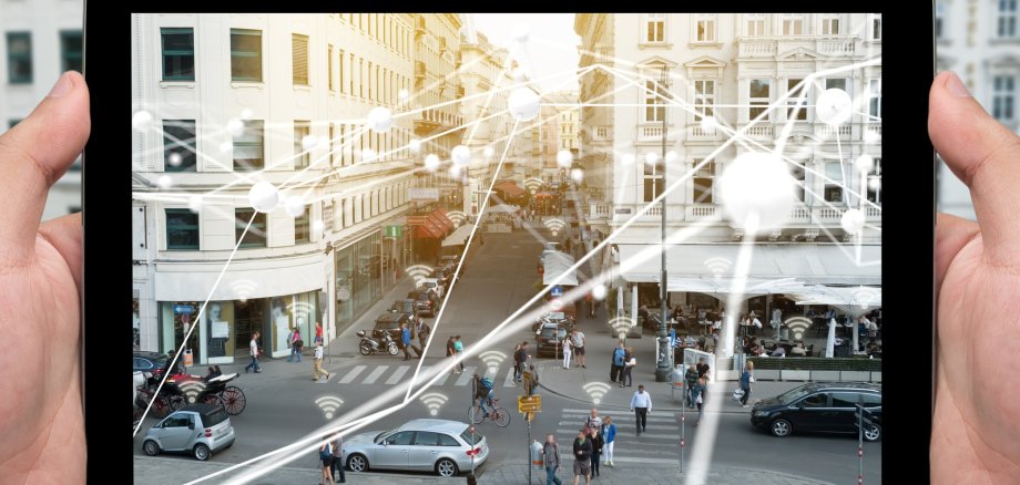 Zwei Hände halten ein Tablet in einer Innenstadt. Blick durch das Tablet auf die digitale Innenstadt.