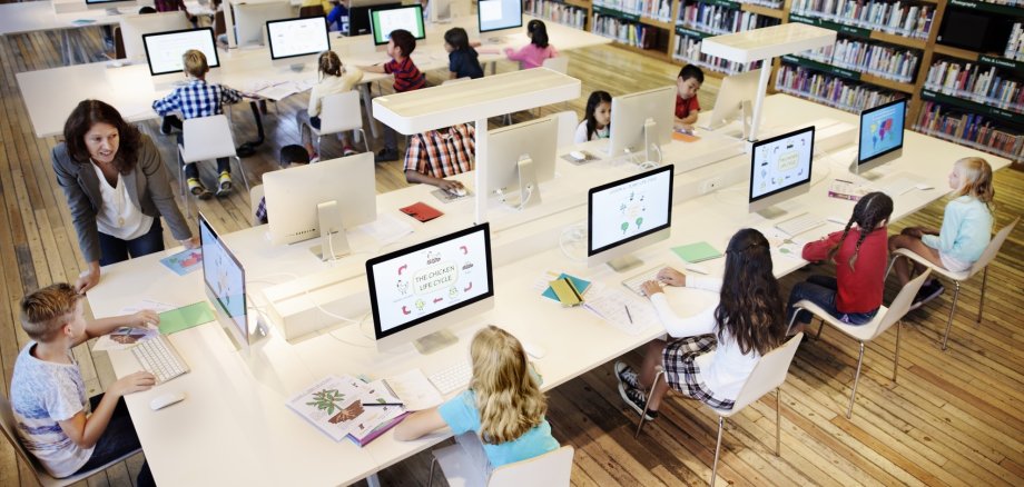 Junge Schüler sitzen an großen Tischen vor jeweils eigenen Rechnern in einer Bibliothek.