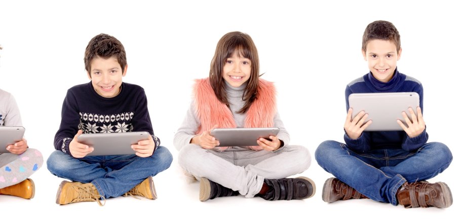 Fünf Schüler - drei Jungen und zwei Mädchen - sitzen im Schneidersitz nebeneinander auf dem Boden und halten jeweils ein Tablet in der Hand. Der Rest des Bildes ist weiß.