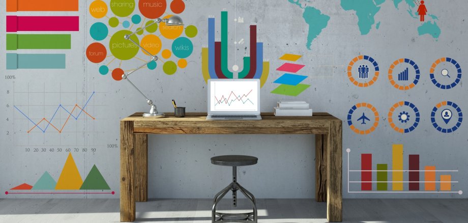 Ein Schreibtisch steht an einer grauen Wand. Auf dem Tisch steht ein Laptop und davor ein Hocker. Auf der Wand sind eine Vielzahl von Grafiken abgebildet.