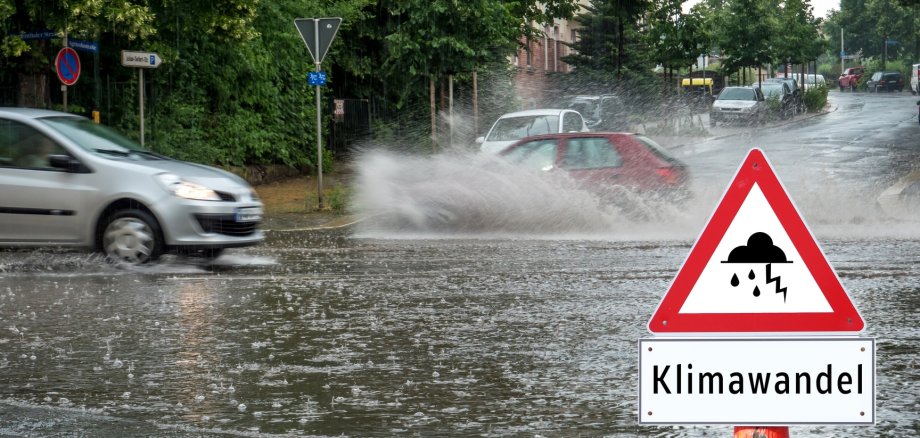 Ein Auto fährt durch eine überflutete Straße und vorne steht das Schild mit der Aufschrift "Klimawandel". Ein Auto fährt durch eine überflutete Straße und vorne steht das Schild mit der Aufschrift "Klimawandel".
