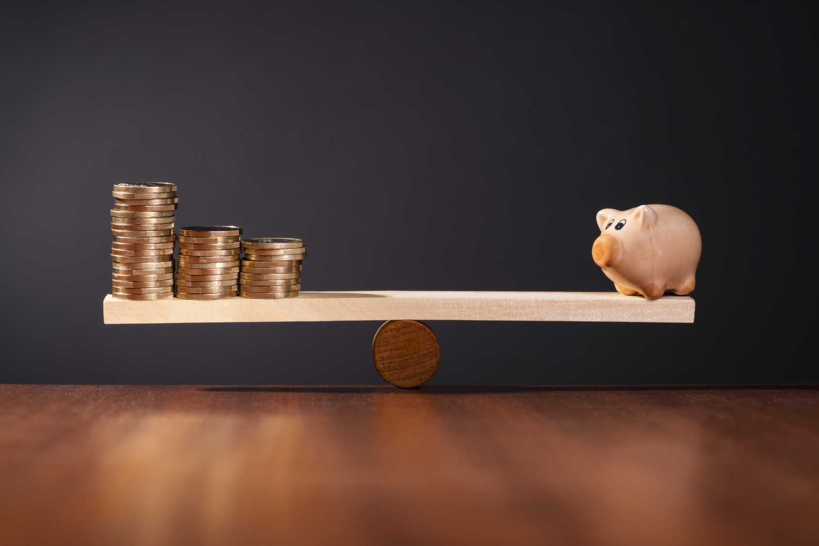 Wippe mit Sparschwein auf der einen Seite und Stapel mit Geld auf der anderen Seite.