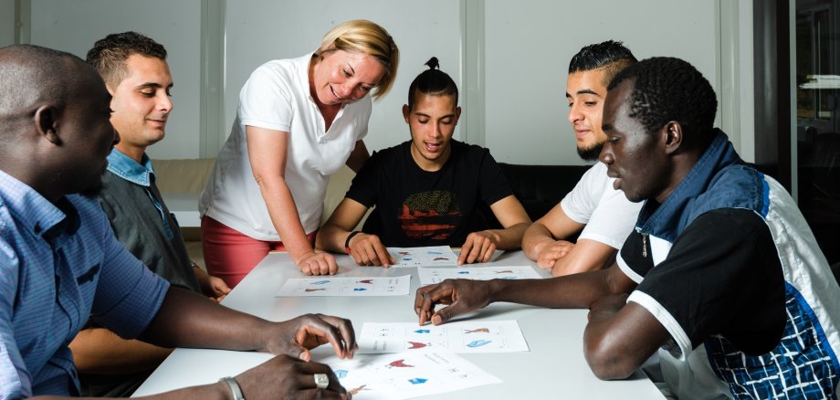 An einem Tisch sitzen fünf Flüchtlinge die eine Lerngruppe bilden. Am oberen Ende erklärt die Lehrerin einem Teilnehmer etwas