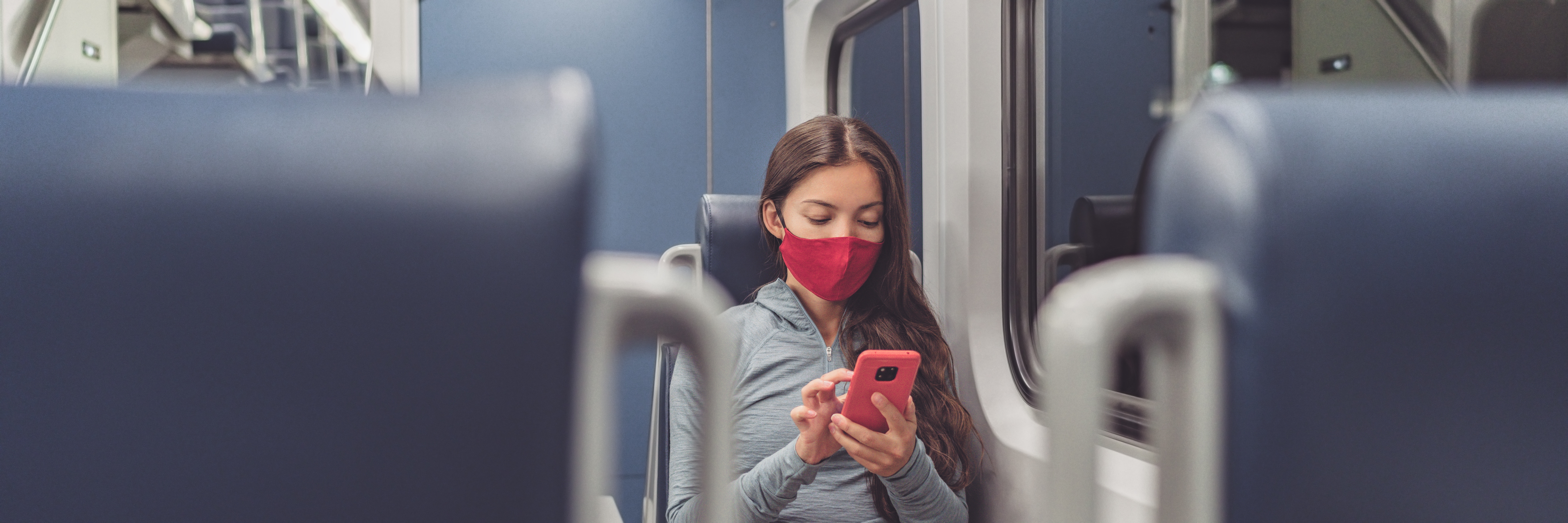 Eine Frau sitzt im Zug. Sie trägt dabei eine Maske und schaut auf ihr Smartphone.  Eine Frau sitzt im Zug. Sie trägt dabei eine Maske und schaut auf ihr Smartphone.