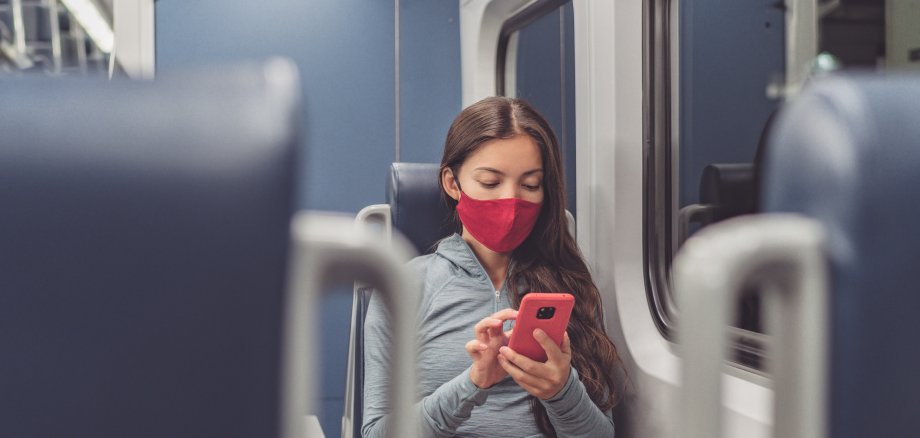 Eine Frau sitzt im Zug. Sie trägt dabei eine Maske und schaut auf ihr Smartphone. 