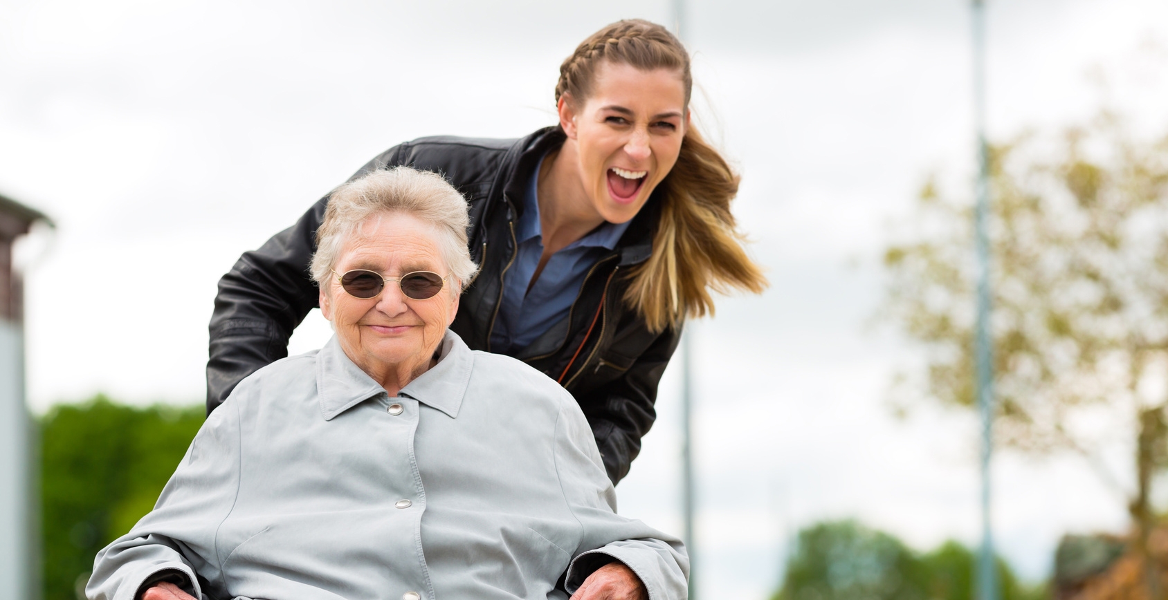 Eine junge Frau, die eine ältere Frau im Rollstuhl anschiebt und lacht. Eine junge Frau, die eine ältere Frau im Rollstuhl anschiebt und lacht.