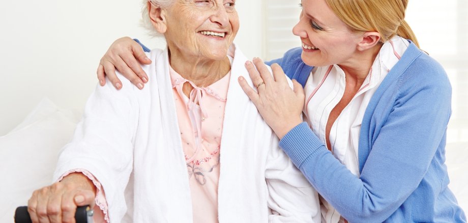 Lachende Seniorin schaut eine junge Frau an, die die Hände um ihre Schultern legt.