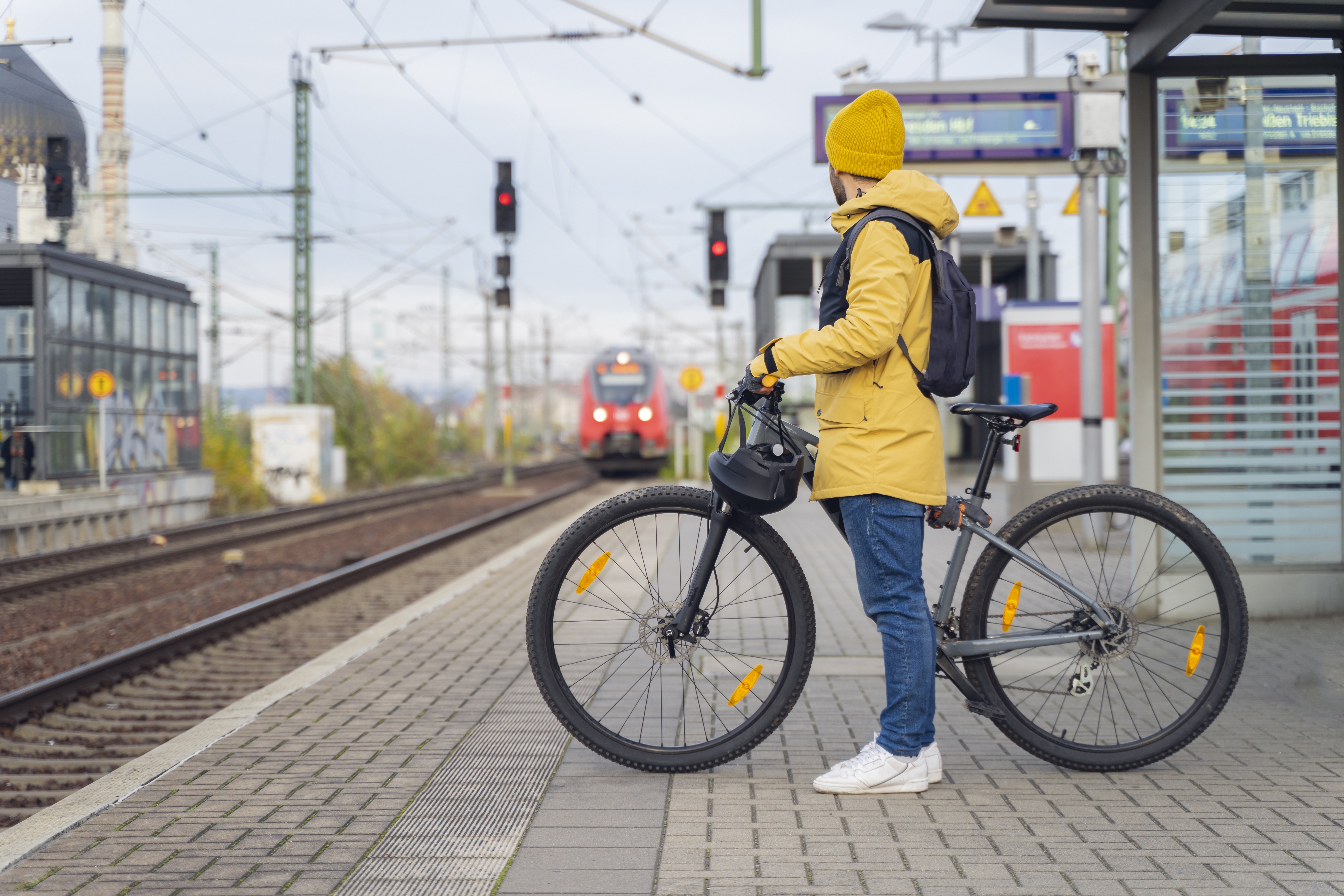 Ein Mann in einer gelber Wetterjacke und mit gelber Mütze wartet mit seinem Fahrrad auf den Zug. Im Hintergrund ist der ankommende Zug zu sehen. Ein Mann in einer gelber Wetterjacke und mit gelber Mütze wartet mit seinem Fahrrad auf den Zug. Im Hintergrund ist der ankommende Zug zu sehen.