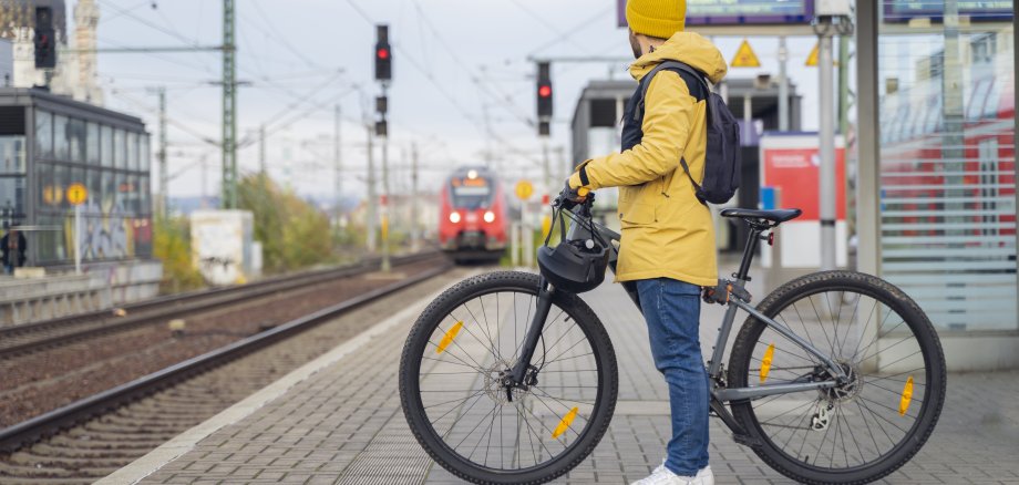 Ein Mann in einer gelber Wetterjacke und mit gelber Mütze wartet mit seinem Fahrrad auf den Zug. Im Hintergrund ist der ankommende Zug zu sehen.