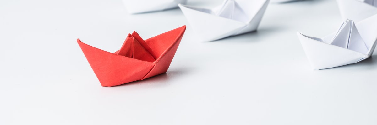 Ein Dreieck aus weißen Papierbooten an dessen Spitze ein rotes Papierboot steht. Ein Dreieck aus weißen Papierbooten an dessen Spitze ein rotes Papierboot steht.