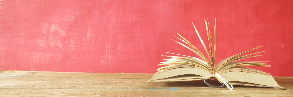 Aufgeschlagenes Buch liegt auf Holztisch vor einer roten Wand. Aufgeschlagenes Buch liegt auf Holztisch vor einer roten Wand.