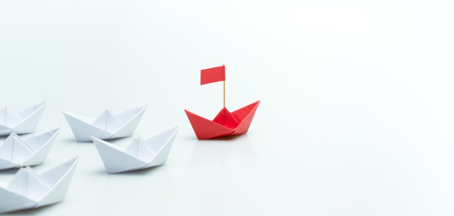 Drei Eck aus weißen Papierbooten, an dessen Spitze ein rotes Papierboot steht.