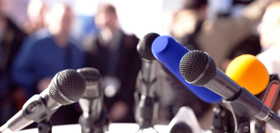 Im Vordergrund stehen zahlreiche nach vorne gerichtete Mikros auf einem Tisch. Im Hintergrund sitzen Menschen. Symbolbild: Pressekonferenz