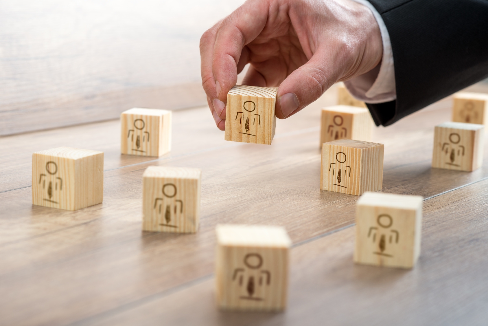 Zahlreiche Holzwürfel mit aufgezeichneten Menschen auf der Vorderseite liegen verteilt auf einem Tisch. Eine Hand hält mit den Fingern einen Wüfel in der Luft.
