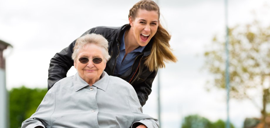Eine junge Frau, die eine ältere Frau im Rollstuhl anschiebt und lacht.