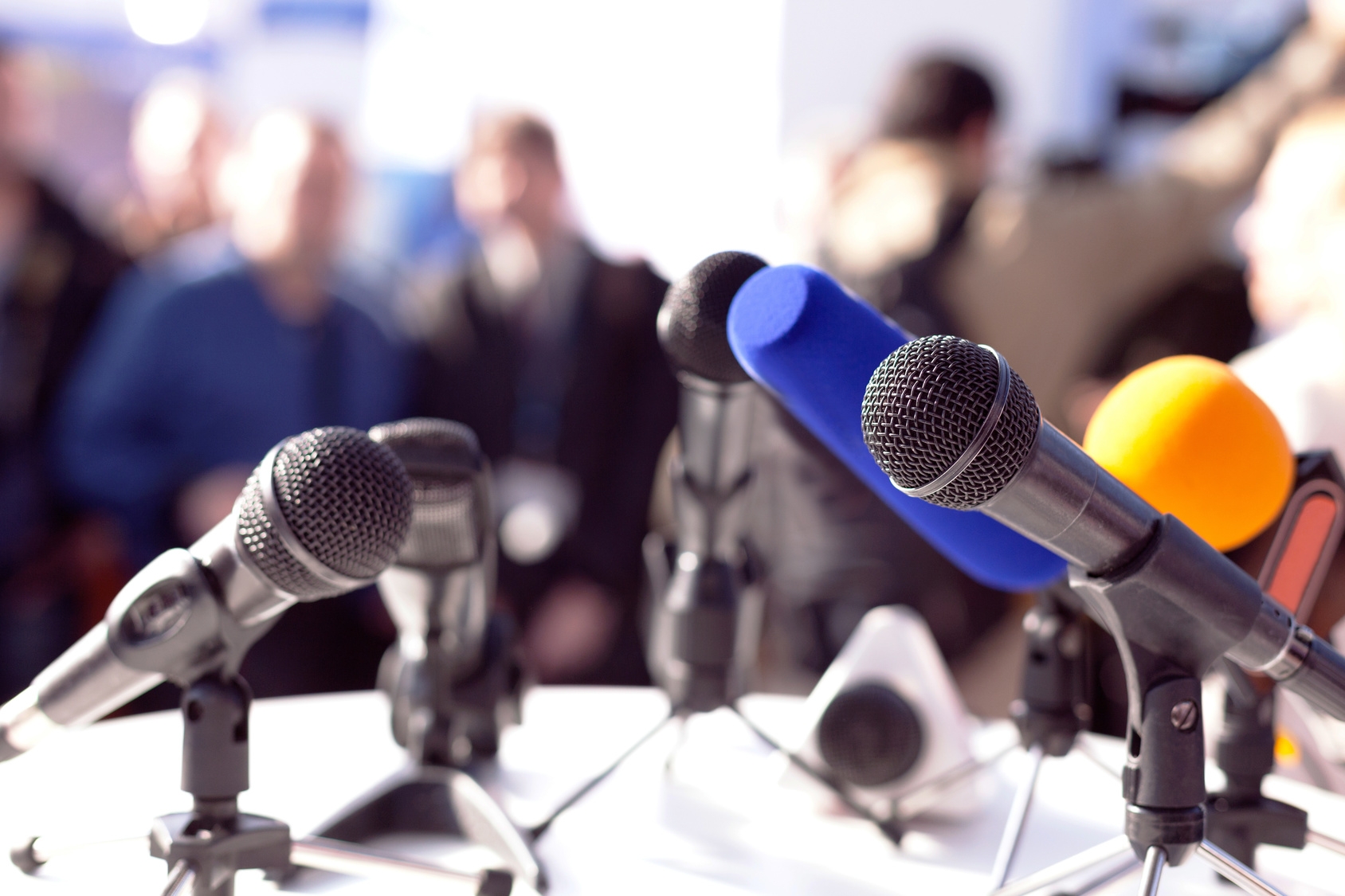 Im Vordergrund stehen zahlreiche nach vorne gerichtete Mikros auf einem Tisch. Im Hintergrund sitzen Menschen. Symbolbild: Pressekonferenz Im Vordergrund stehen zahlreiche nach vorne gerichtete Mikros auf einem Tisch. Im Hintergrund sitzen Menschen. Symbolbild: Pressekonferenz