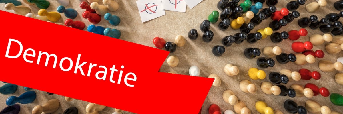 Vogelperspektive auf bunte Holzspielfiguren, welche um Blätter mit angekreuzten Kreisen stehen. Symbolbild für Wahlen. Im Vordergrund ein roter Balken mit der Aufschrift "Demokratie". Vogelperspektive auf bunte Holzspielfiguren, welche um Blätter mit angekreuzten Kreisen stehen. Symbolbild für Wahlen. Im Vordergrund ein roter Balken mit der Aufschrift "Demokratie".
