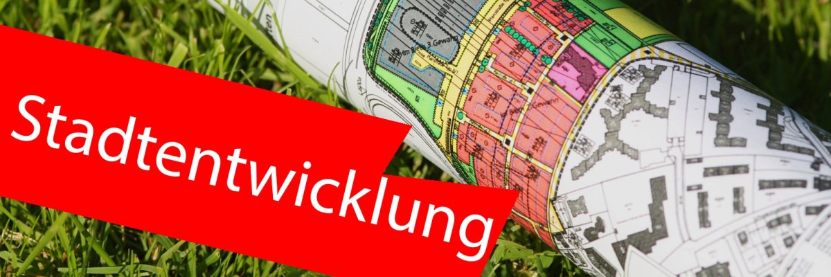Städtebauskizze/Karte zusammengerollt auf Gras. Im Vordergrund ein roter Balken mit der Aufschrift "Stadtentwicklung". Städtebauskizze/Karte zusammengerollt auf Gras. Im Vordergrund ein roter Balken mit der Aufschrift "Stadtentwicklung".