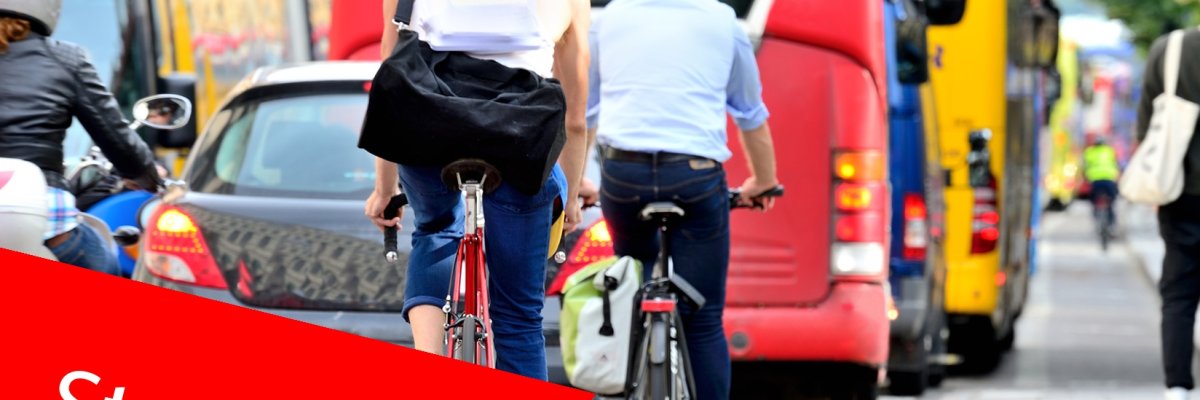 Fahrradfahrer:innen und Autos im Verkehr. Im Vordergrund ein roter Balken mit der Aufschrift "Straßenverkehr". Fahrradfahrer:innen und Autos im Verkehr. Im Vordergrund ein roter Balken mit der Aufschrift "Straßenverkehr".